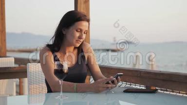 年轻的布鲁内特在日落时坐在海边的咖啡馆里，用她的智能手机端着一杯红酒。